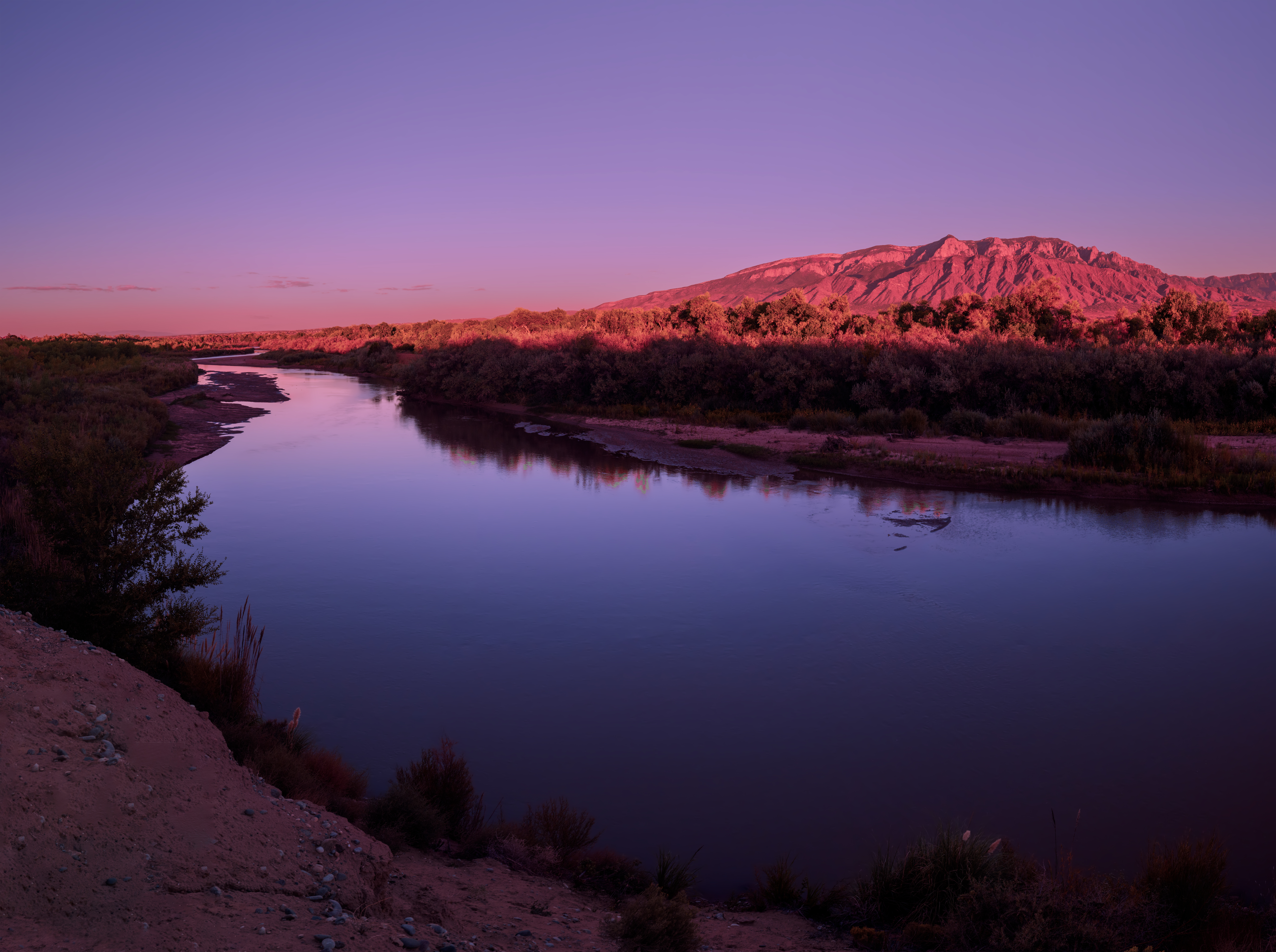 Serene dusk shot of the Rio Grande meandering through brushy banks, reflecting a violet-pink sky with the distant, sunlit mountains behind.