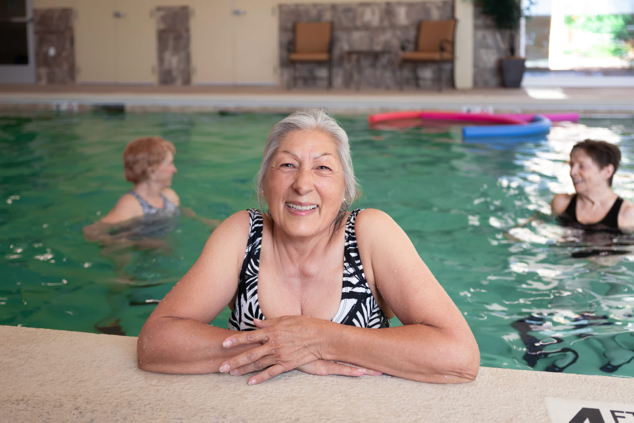An elderly woman leaning on the edge of an indoor pool smiling as two other women exercise in the water behind her.
