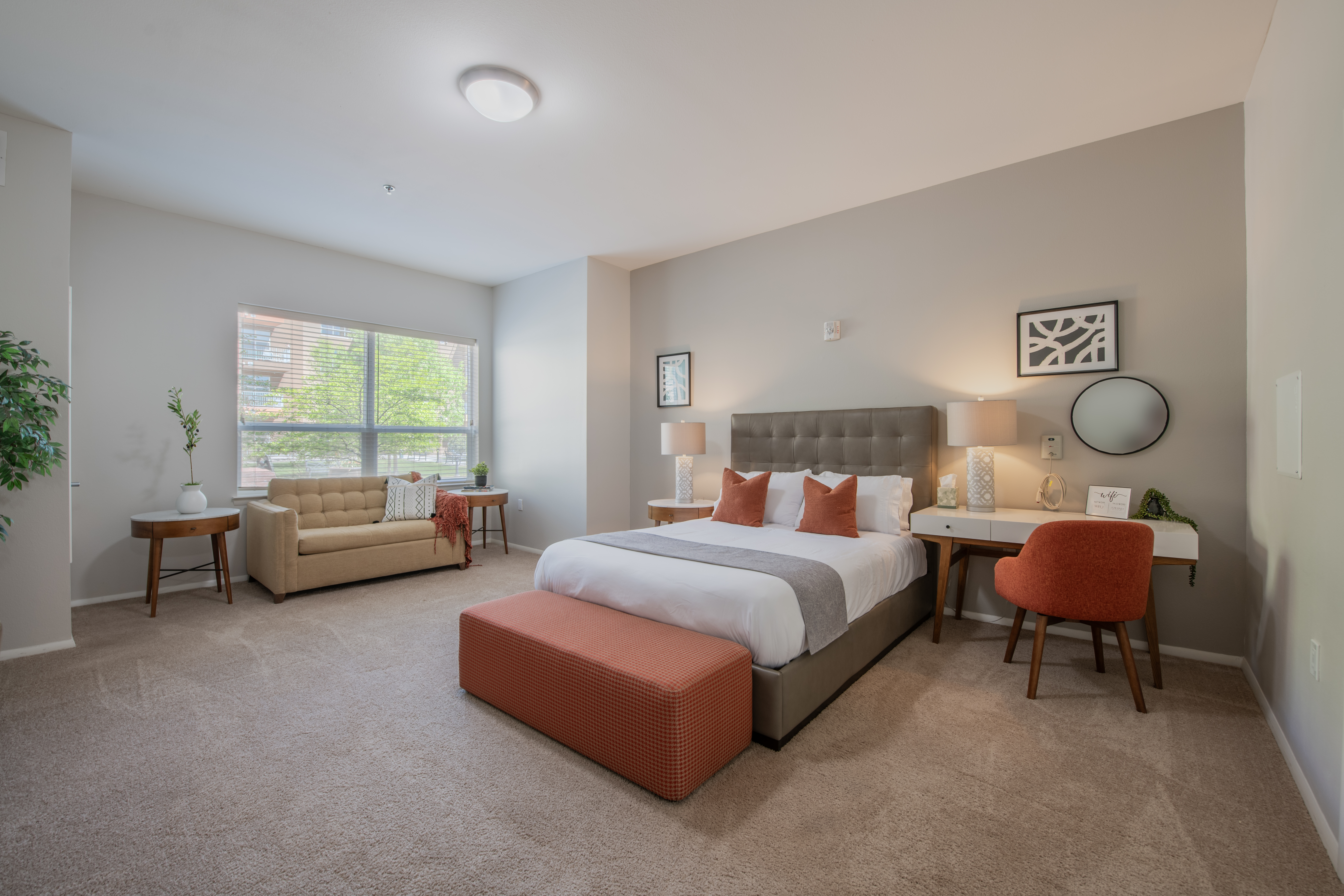 Bright apartment bedroom with upholstered bed, sofa, desk, and large window.